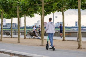 E-scooter rider on sidewalk with trees and pedestrians in the background on a cloudy day