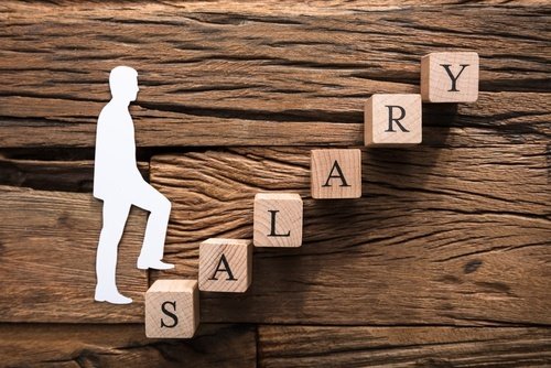 White paper cut-out of a man climbing a stairway of blocks that spell "Salary" against a wooden table