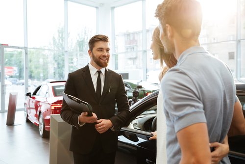 Vendedor de autos en un traje de negocios sosteniendo una tabla de bienvenida a una joven pareja en la sala de exhibición de un concesionario de autos