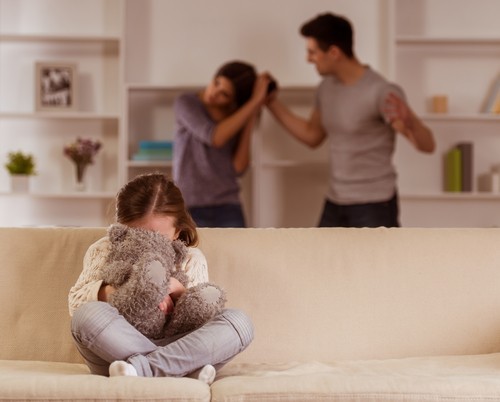 Niña pequeña en un sofá cubriendo su rostro con un oso de peluche mientras un hombre tira del cabello de su madre