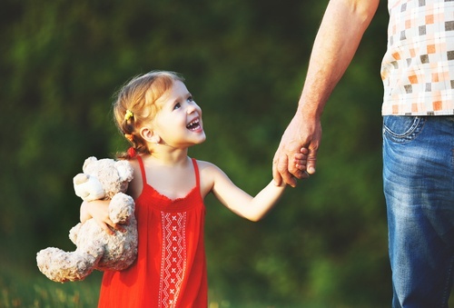 Niña joven con un oso de peluche agarrando la mano de un hombre y mirándolo, sonriendo