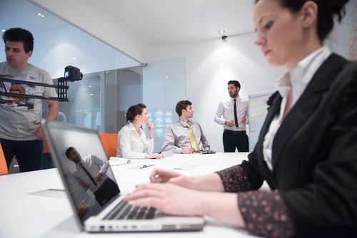 young businesswoman making notes on laptop in conference room with people in background