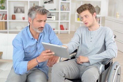 Man with beard holding laptop up to young man in wheelchair