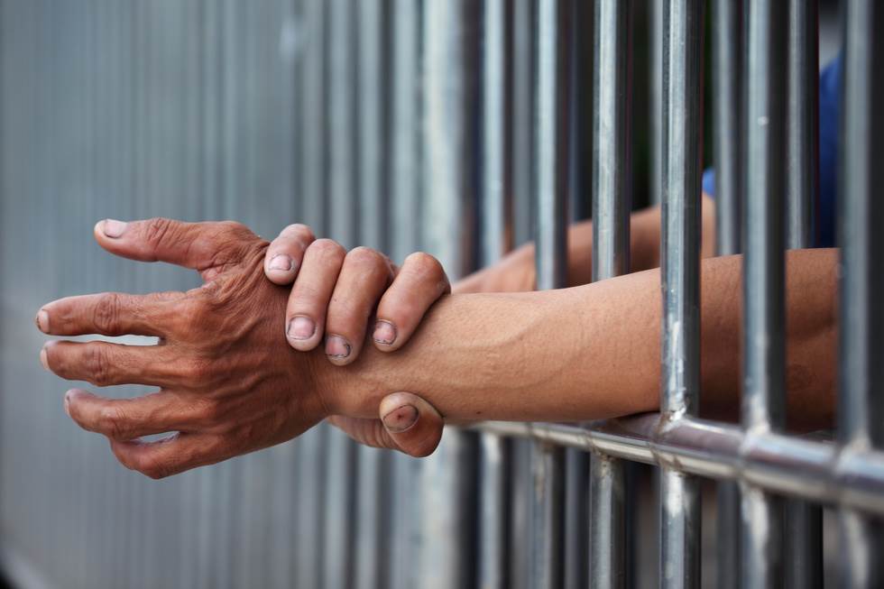 Las manos de los reclusos proyectándose a través de las rejas de la cárcel: una condena por contribuir a la delincuencia de un menor según el Código Penal de California 272 PC puede llevar hasta un año en la cárcel
