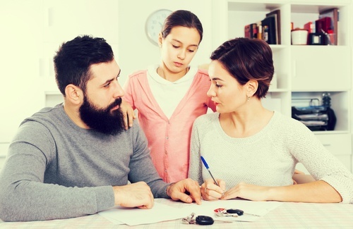 Young girl standing in between parents who are looking unhappy and signing a divorce document.