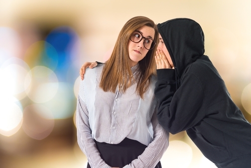 young person in hoodie whispering something into ear of girl wearing button up shirt and glasses