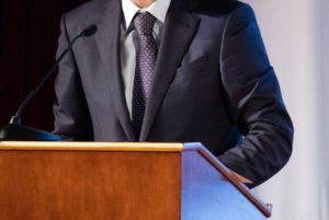 Man in suit speaking behind a podium. 