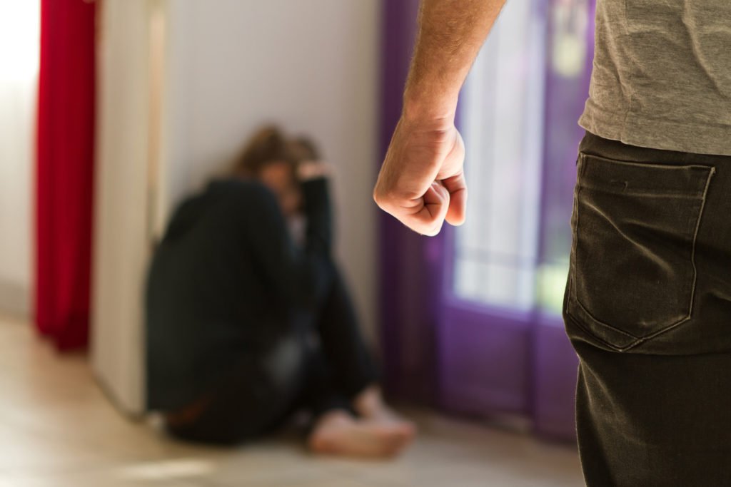 Hombre sosteniendo el puño con su esposa agachada en la esquina para ilustrar la violencia doméstica.