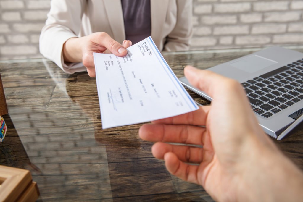Close-up of employer handing check to employee over a glass desk with a laptop