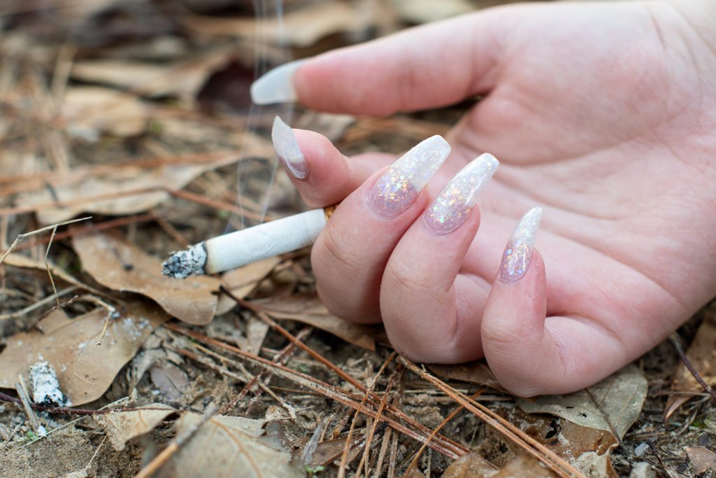 Woman's hand holding lit cigarette inches away from dry leaves as an example of reckless endangerment per CRS 18-3-208