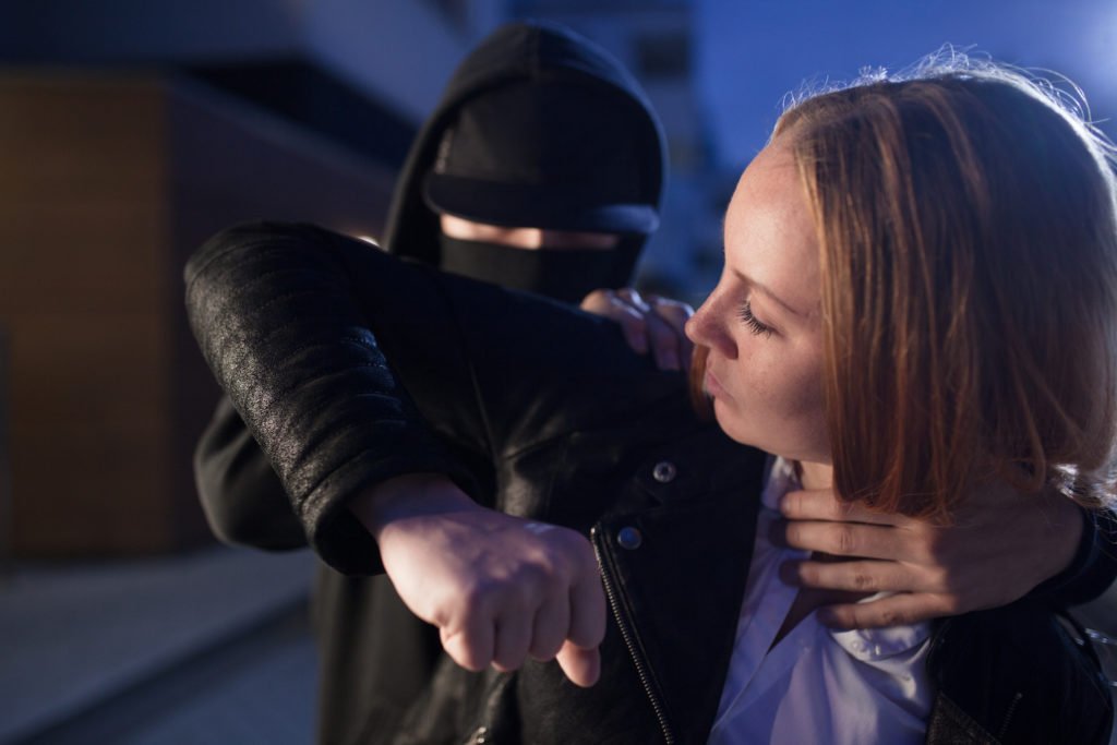 Woman fighting back with her elbow against a masked assailant on street who is holding her neck and shoulders