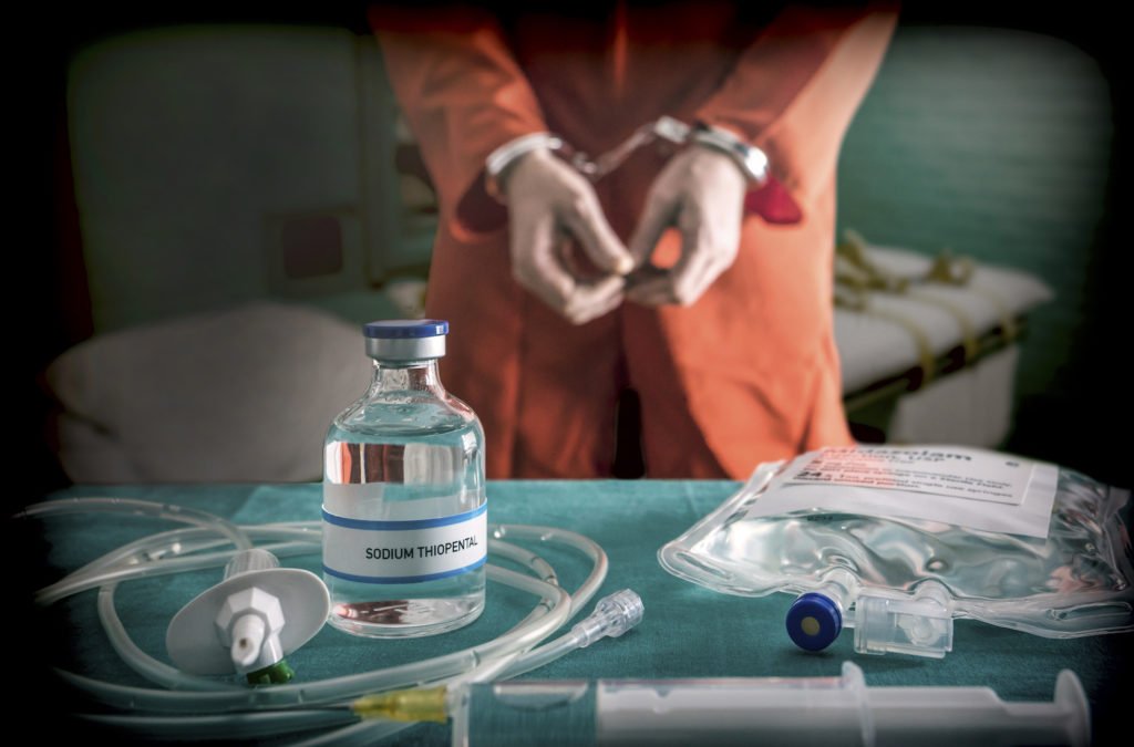 Death row inmate in cuffs in front of table with lethal injection supplies