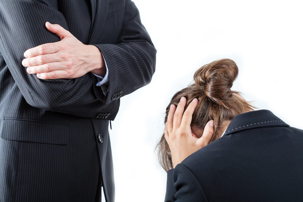 Mujer sentada en la oficina siendo regañada por su jefe masculino de pie sobre ella como ejemplo de acoso laboral, en un fondo blanco