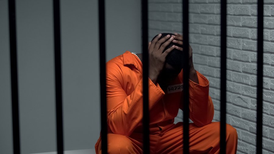 Recluso en traje naranja sosteniendo sus manos y sentado detrás de barras de prisión contra un fondo de ladrillos