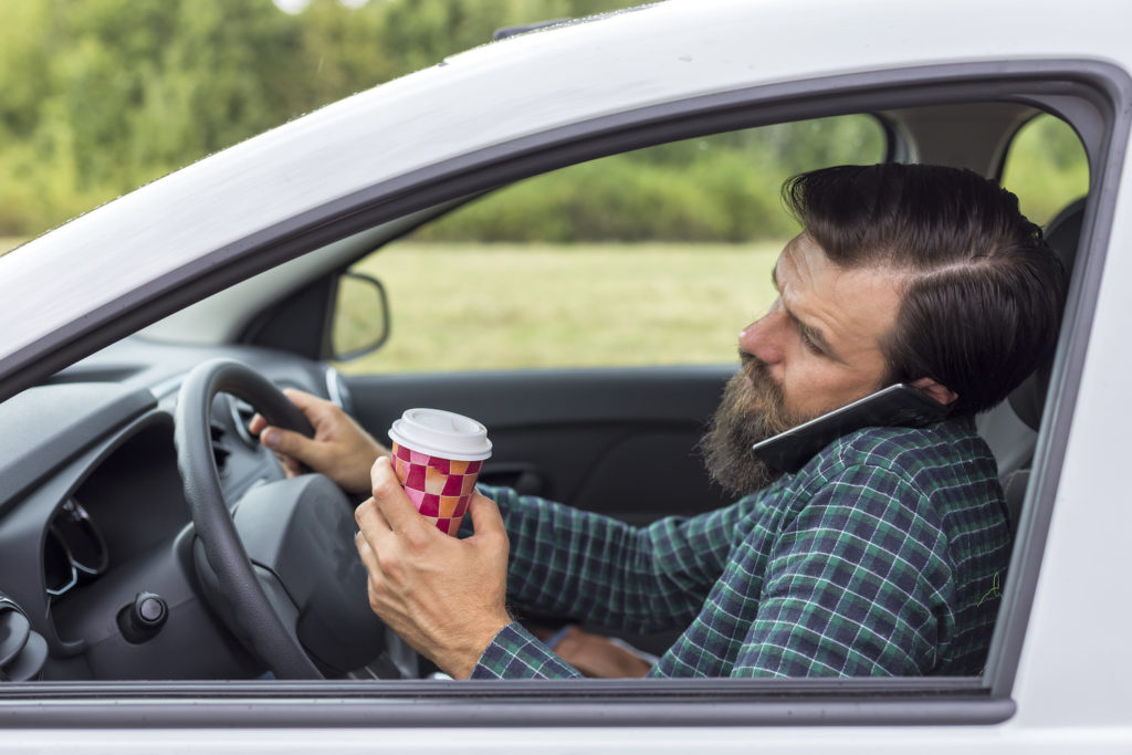 Conductor detrás del volante hablando por teléfono celular contra el oído y sosteniendo café con una mano