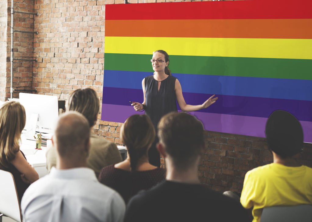 Woman in front of rainbow flag giving speech to office - workplace discrimination based on sexual orientation is illegal in California
