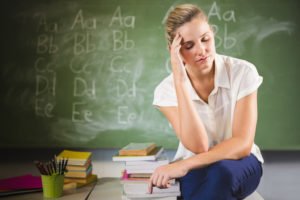 Teacher with headache sitting on desk in class room in front of blackboard