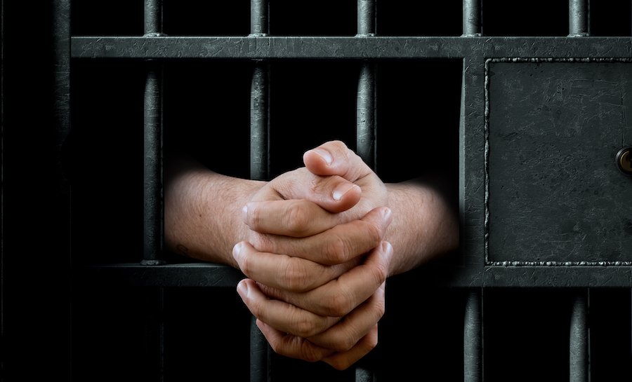 Una imagen cercana de una puerta de celda de prisión con poca luz con brazos extendidos en una posición de oración apretada
