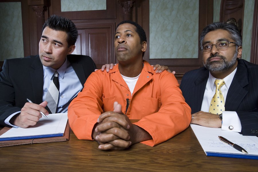 Man in orange jumpsuit flanked by attorneys in court