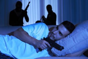 Man in bed with gun with two armed intruders behind him