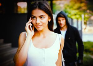 Hombre con capucha acechando a una joven que usa su teléfono en la calle