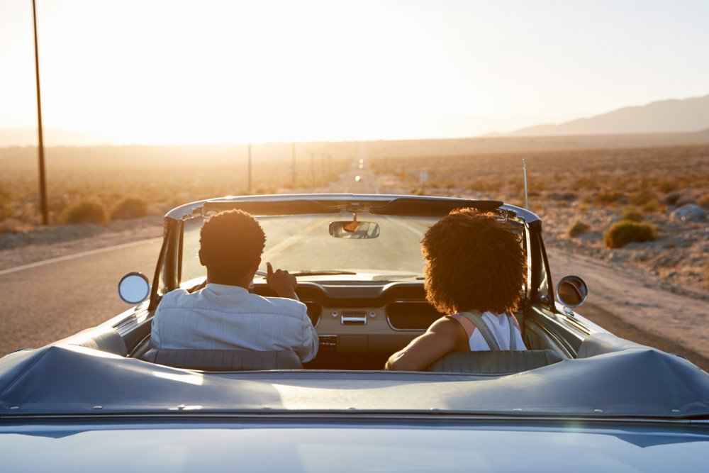 Una pareja conduciendo un convertible hacia el atardecer.