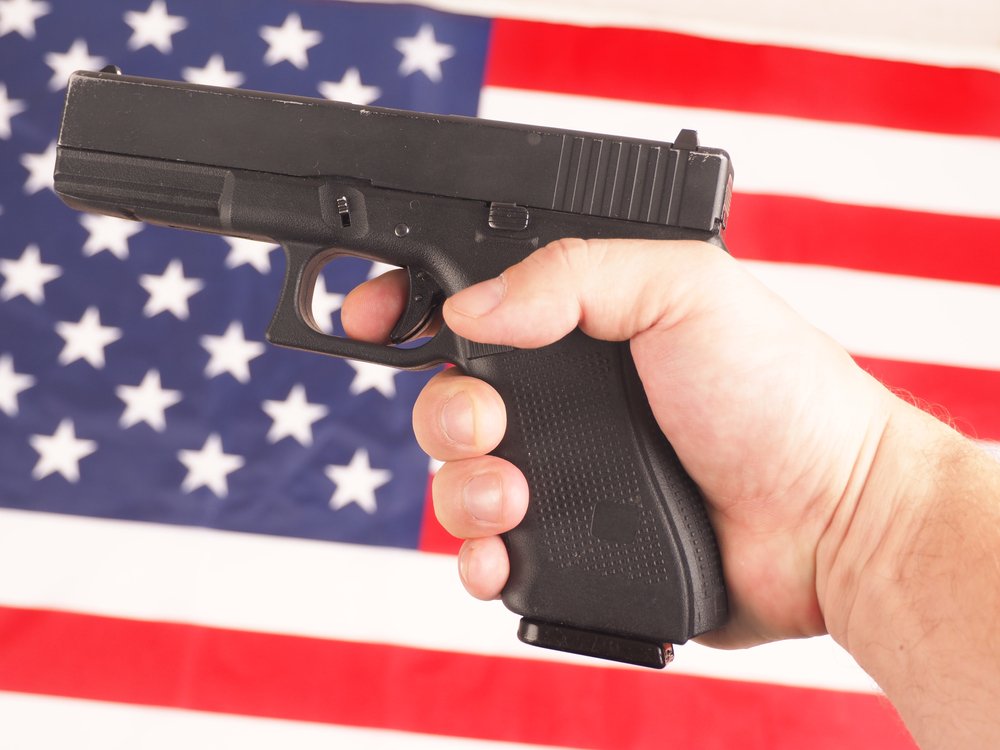 Una mano sosteniendo un arma frente a una bandera estadounidense.