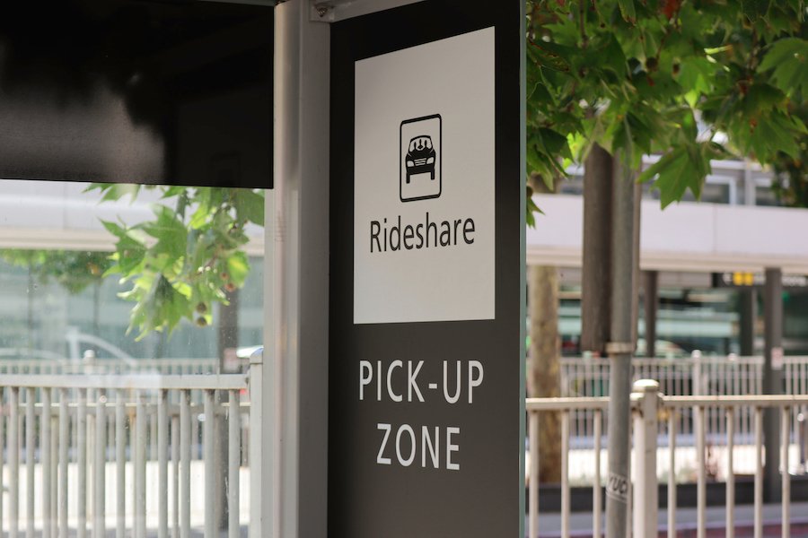 A black and white sign that says "Rideshare Pick-Up Zone" outside of an airport on a sunny day