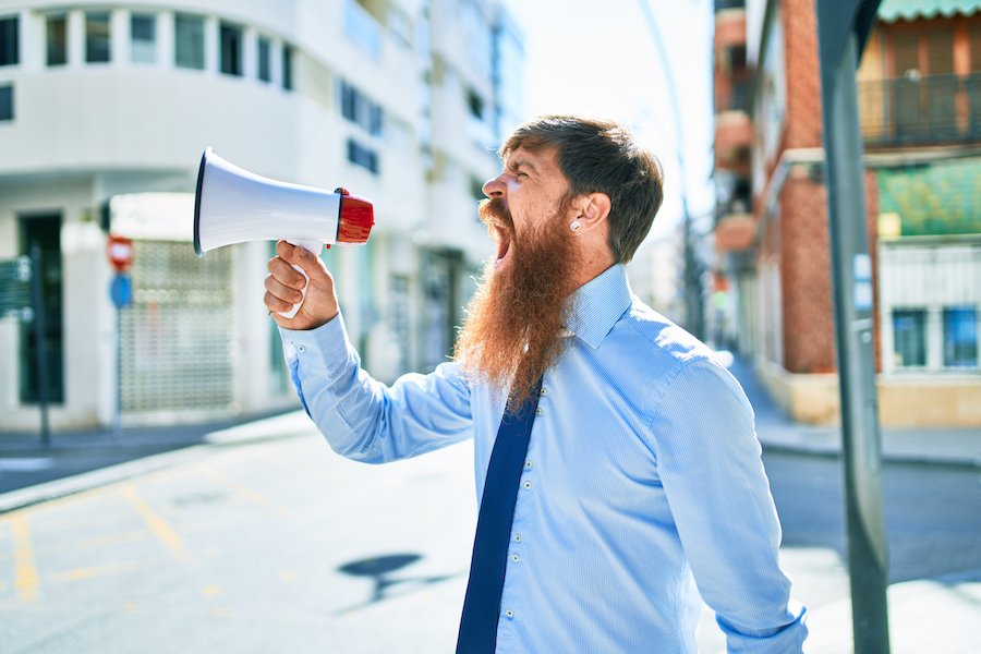 Young redhead businessman with angry expression. Screaming using megaphone at street of city.