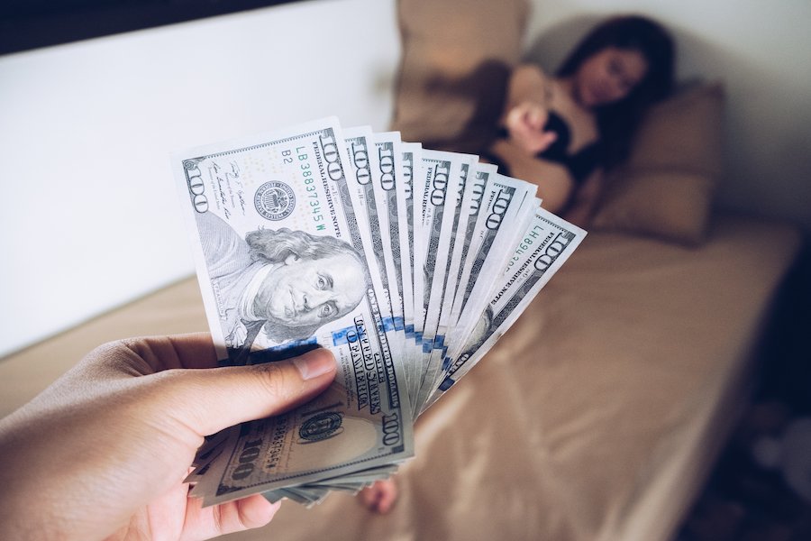 John holding cash while and a prostitute lies on a bed