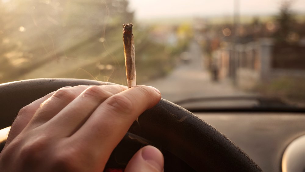 Person with a joint in his hand resting on a steering wheel as an example of a Vehicle Code 23221 VC violation