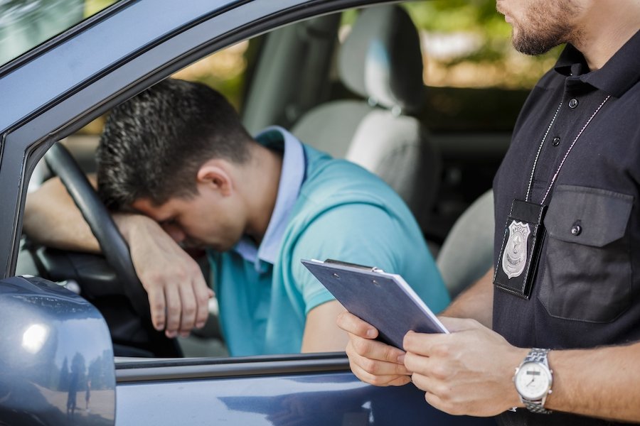 Una toma de un joven conductor manteniendo su cabeza en un volante y un policía de pie junto al coche
