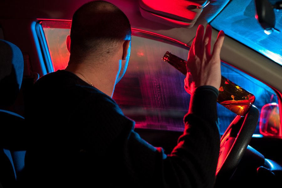 Drunk driver sitting in car with alcohol bottle and looking through the window at police car.