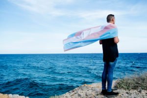 Person wearing transgender flag as cape against the ocean
