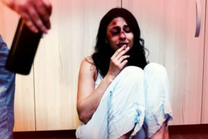 A dejected woman sitting on the ground, with a bloody face, smoking a cigarette.