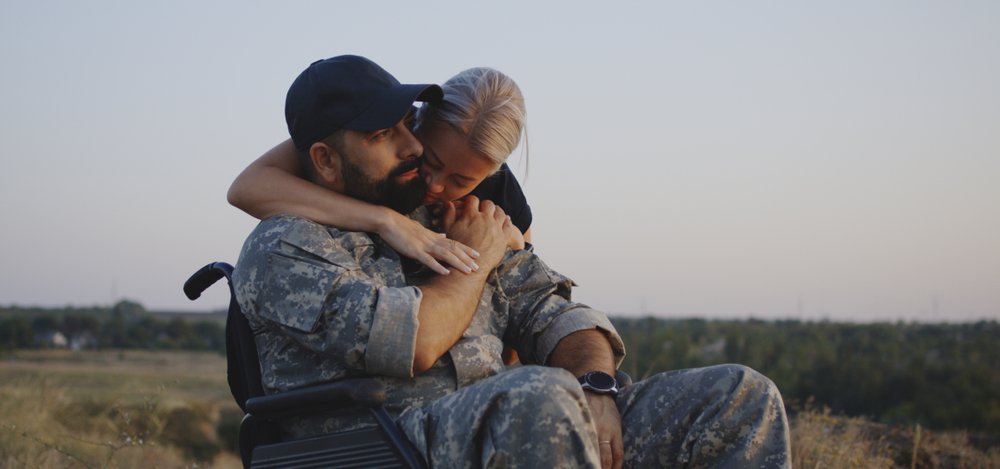 A disabled veteran in a wheelchair being hugged from behind by his wife.