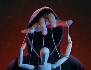 A hand coming out of a hooded sweater holding the dangling lines of a puppet, against a menacing red background.