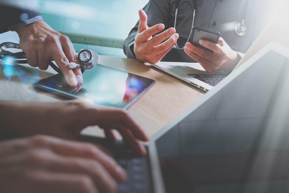 Closeup of a physician meeting with their hands touching a stethoscope, and iPad, a laptop, and an upright computer