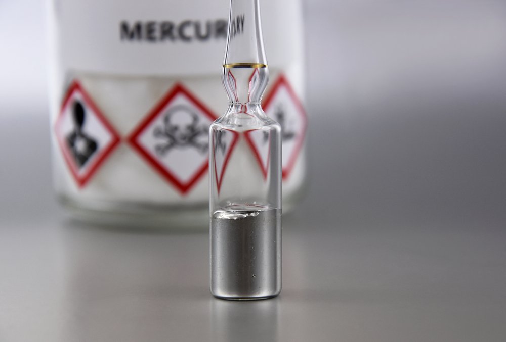 A medical canister holding liquid mercury, in front of poison warning signs.