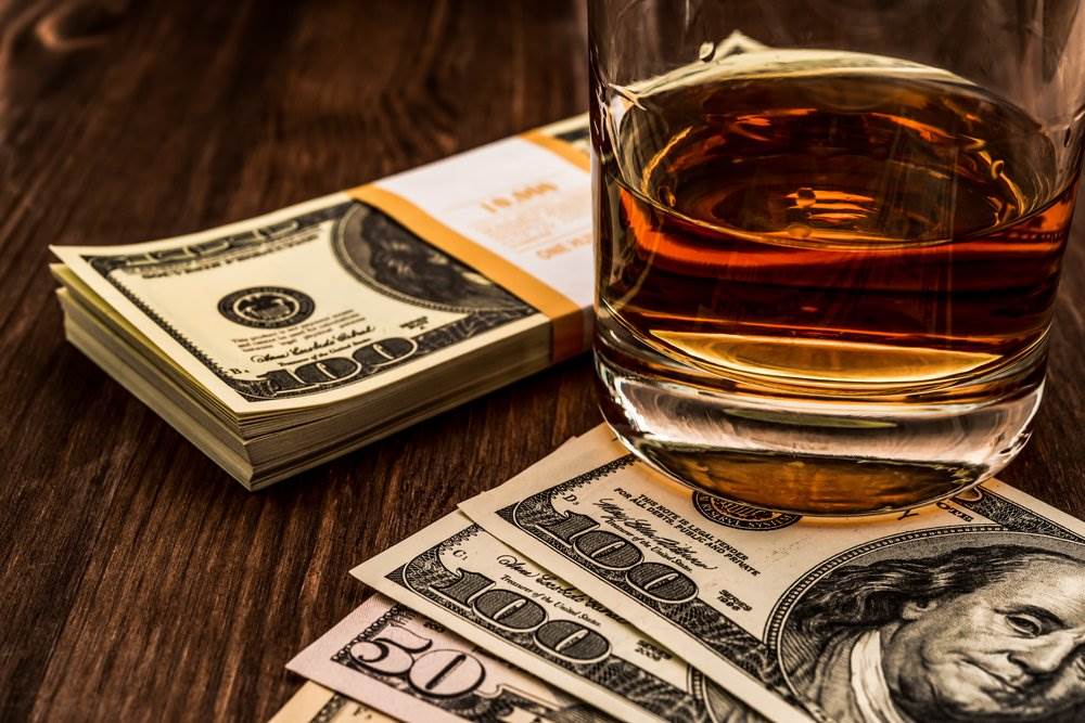 A stack of one hundred dollar bills next to a glass of whiskey. A DUI can be very costly.