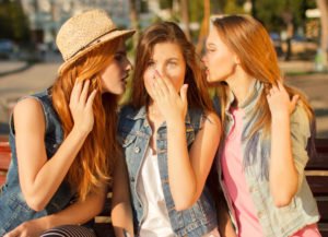 Two women speaking into the ear of a surprised third woman.