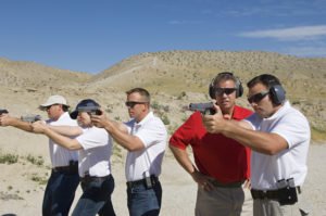 A group of men shooting pistols in front of an instructor out in the desert.