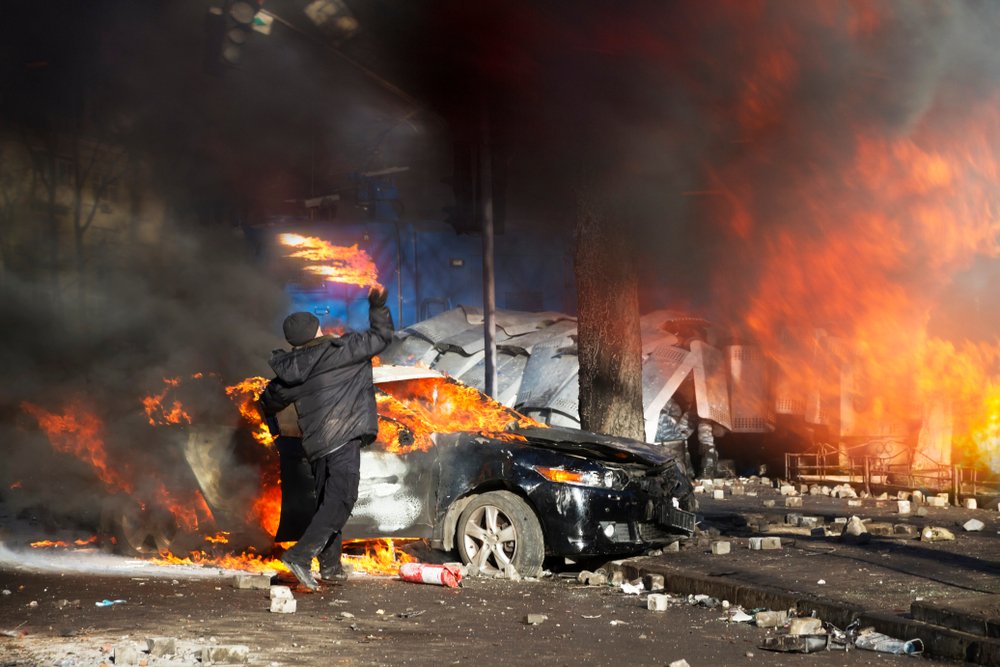 Rioters throwing object behind a burning vehicle.