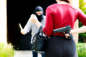 A woman holding a pistol behind her back as a robber approaches her with a knife.
