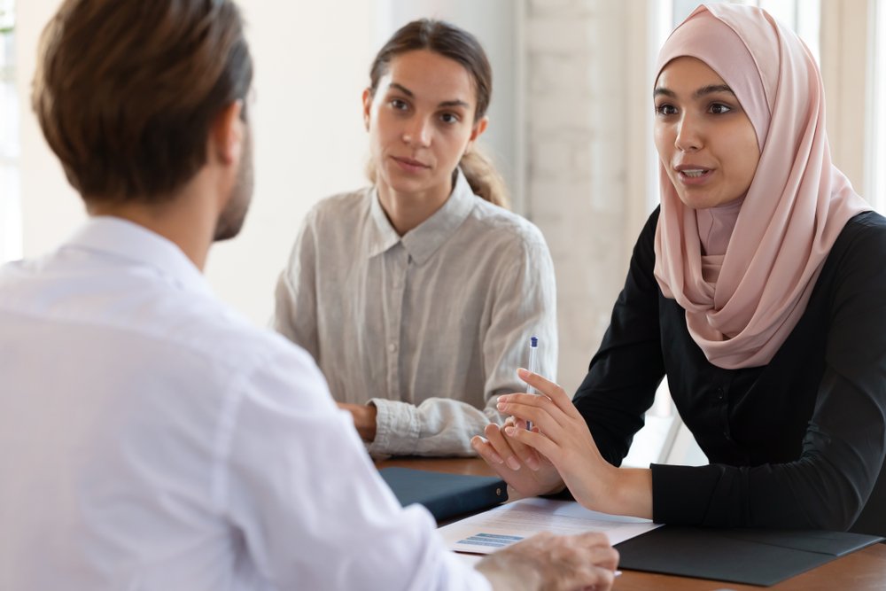 An Asian Muslim lady wearing a Hijab speaking with her Caucasian coworkers.