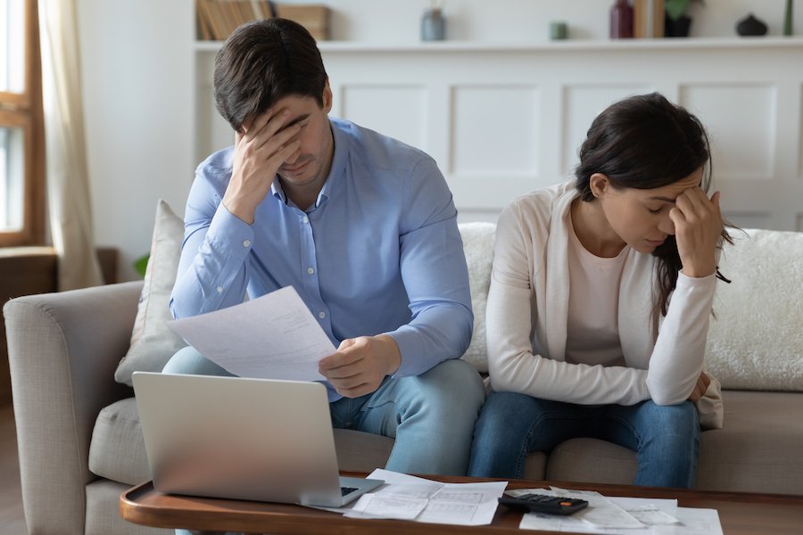 Young couple fretting over unpaid bills while sitting on couch in front of laptop