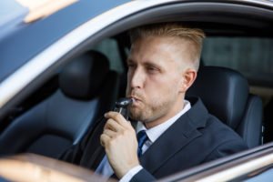 A suited man blowing into a breathalyzer in his car.