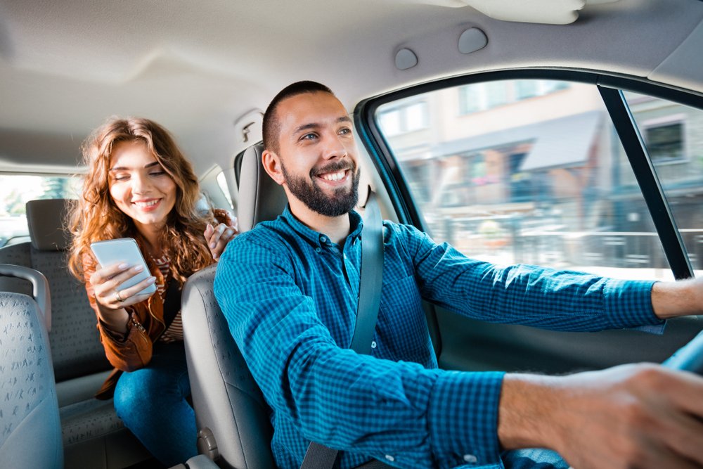 A smiling rideshare driver with an excited passenger behind him.