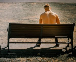 A backside view of a shirtless man sitting on a park bench possibly exposing himself indecently.