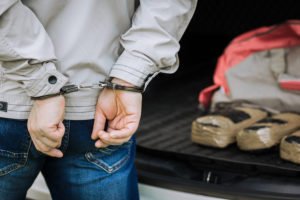 Man in handcuffs by car trunk with packaged drugs